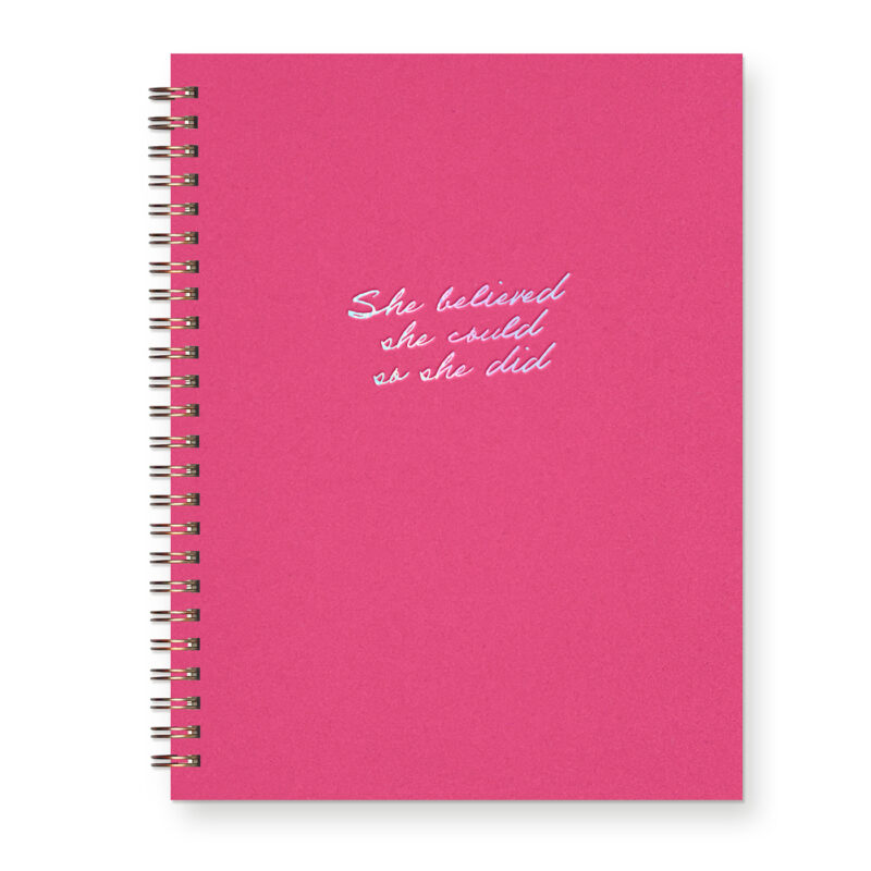 A script font reads "She believed she could so she did" in iridescent foil on a hot pink journal