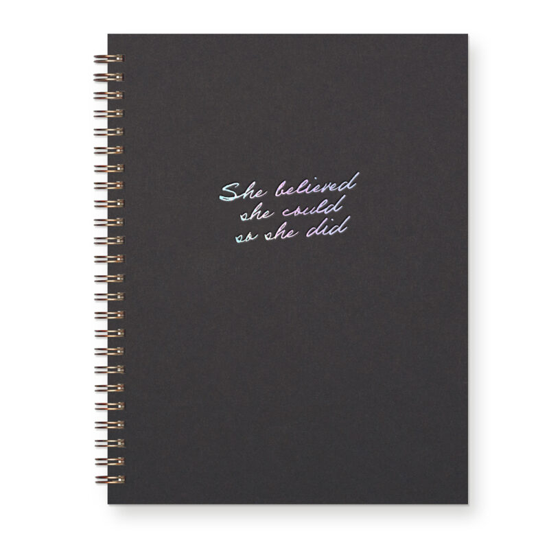 A script font reads "She believed she could so she did" in iridescent foil on a black journal